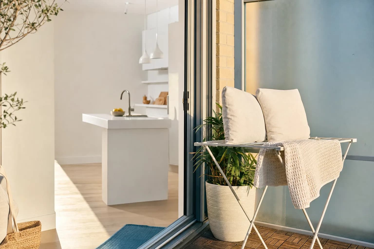 A sunny Toronto balcony scene. Two couch cushions and a folded bath mat sunning on a drying rack, a spotless white kitchen visible through the open sliding door, a small bowl of lemons on the counter, bright July light.